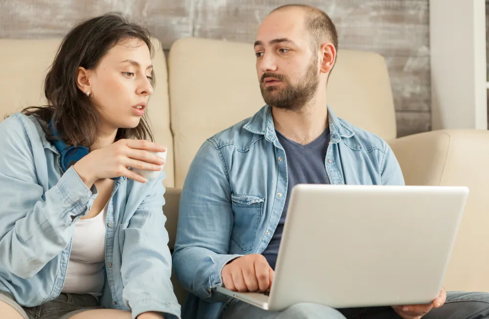 Young couple sat together in a living room with a laptop, illustrating an online relationship counselling session attended jointly from home.