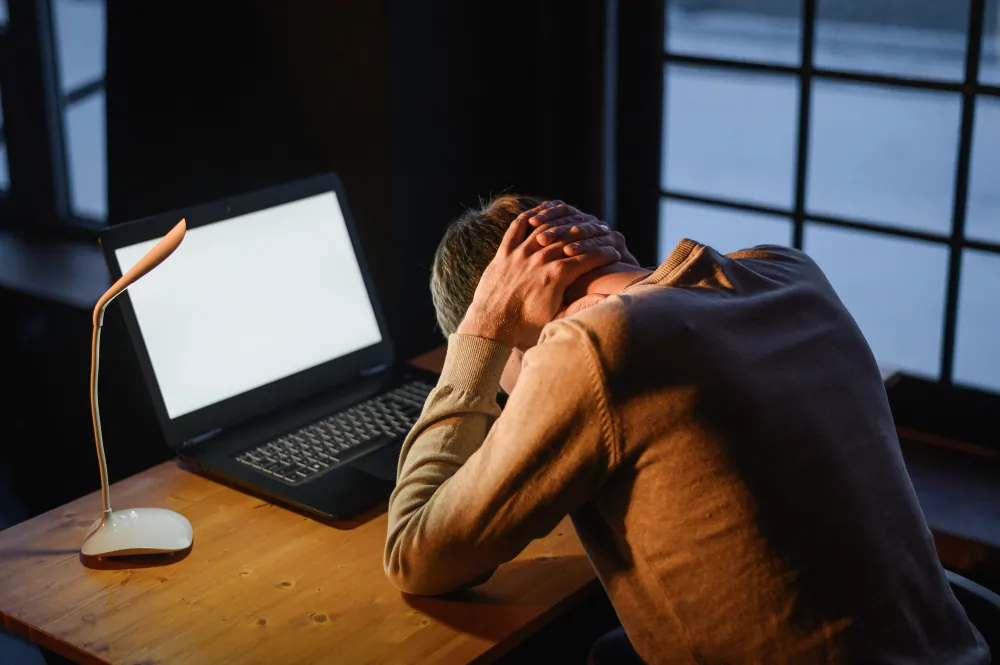 An exhausted man slumped over his desk in low light beside a glowing laptop, illustrating autistic burnout in adults.