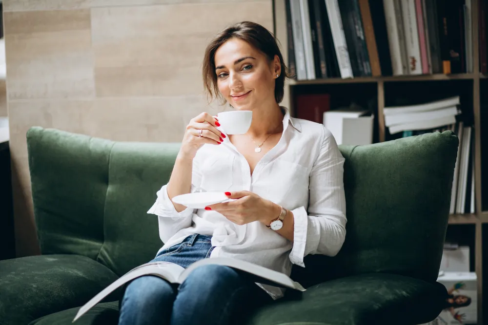 Woman sitting on a sofa reading a book about changing thought patterns, reflecting calmly with a cup of tea, illustrating the self-guided practice of cognitive reframing