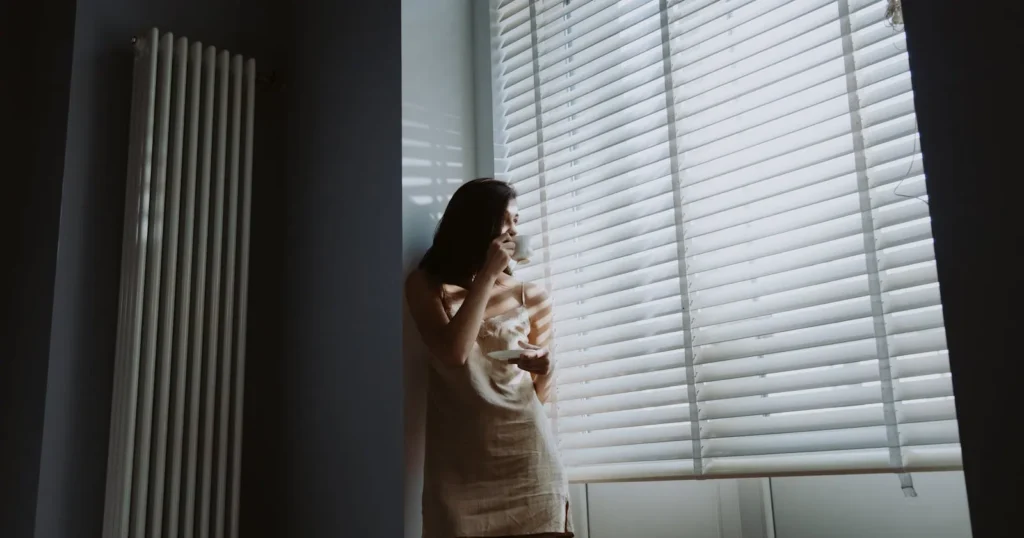 A woman standing by a window in soft natural light, holding a cup during a quiet, reflective moment