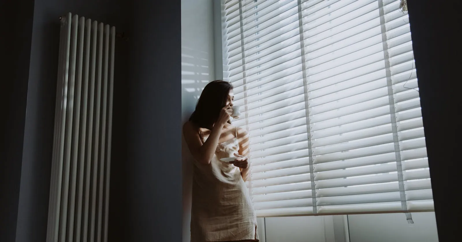 A woman standing by a window in soft natural light, holding a cup during a quiet, reflective moment