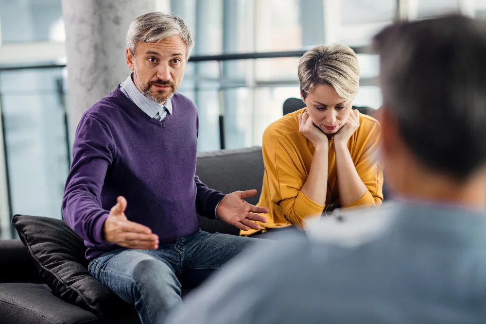 A couple sitting on a sofa in a calm therapy room, mid-conversation with a therapist, weighing up whether couples therapy works