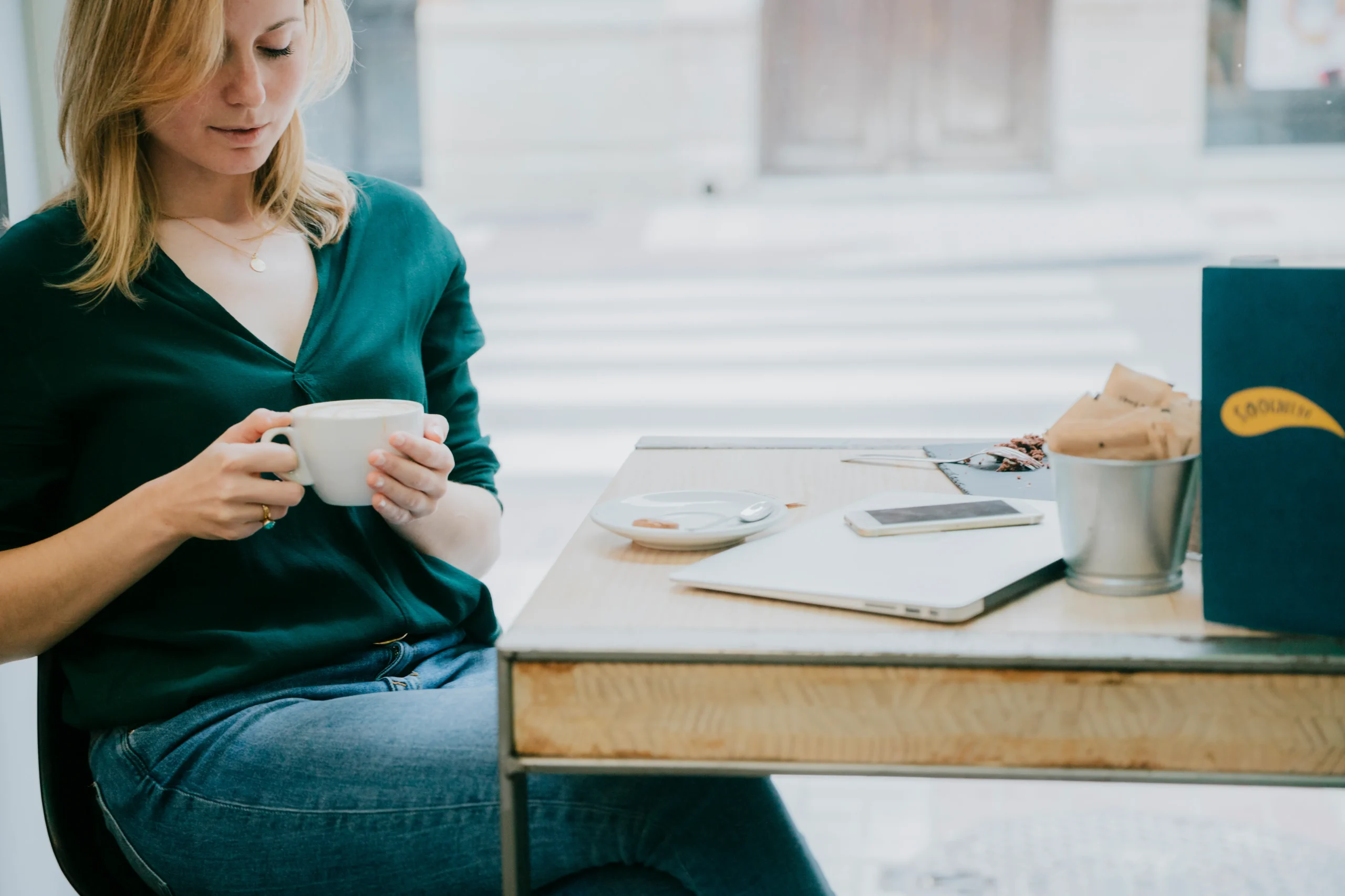 A woman pauses with a cup of coffee at a cafe table, laptop closed beside her